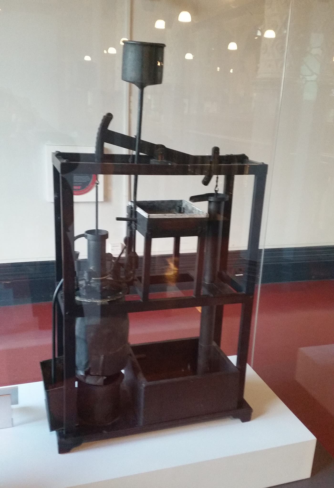 格拉斯哥大学亨特博物馆展柜里的纽科门机模型：深色框架，顶上一根两头带弧形头的横梁，左下是锅炉和气缸，右边是一根竖直的泵管