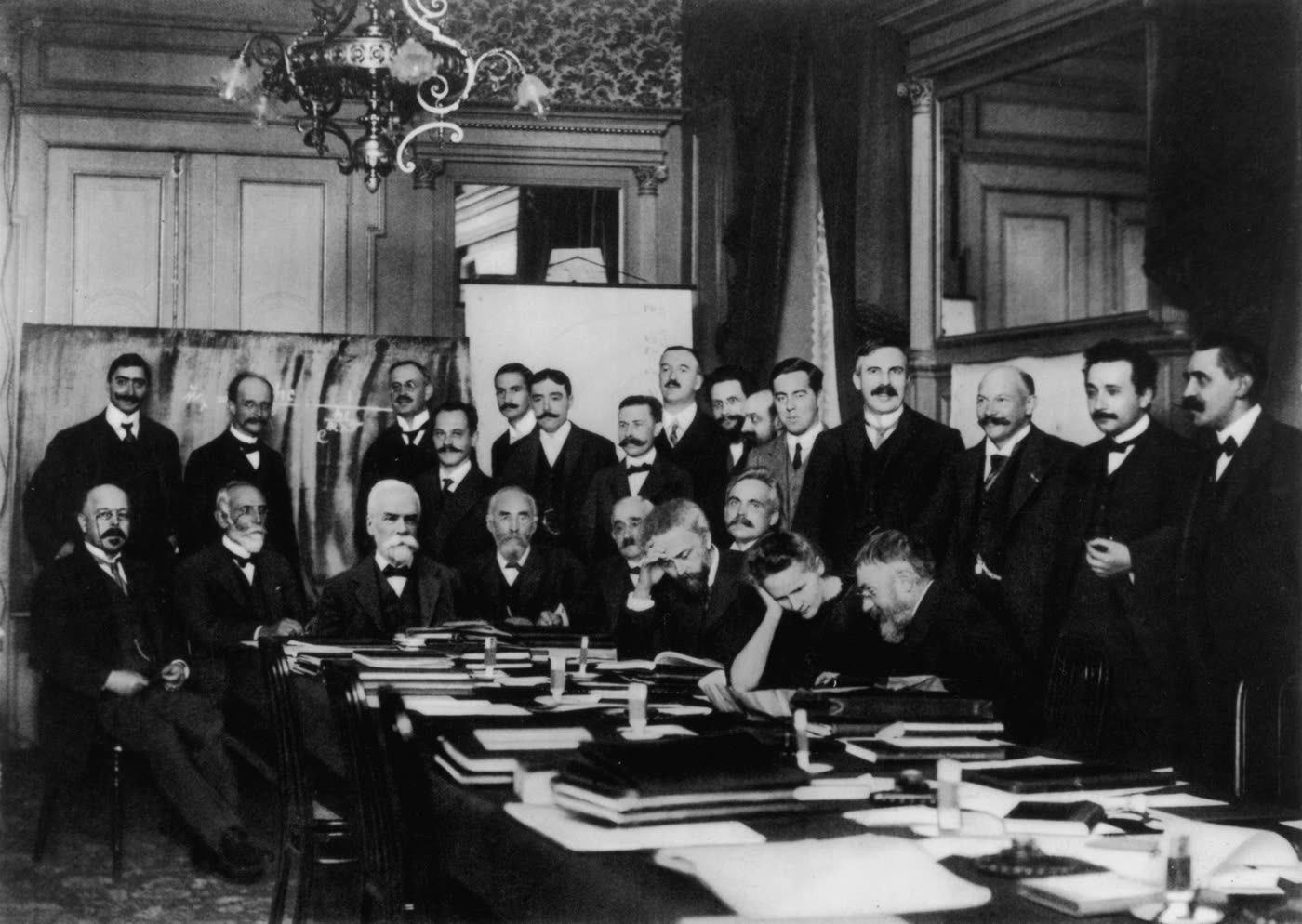 1911 年第一届索尔维会议合影，二十多人围着一张堆满文件的长桌，有人坐有人站